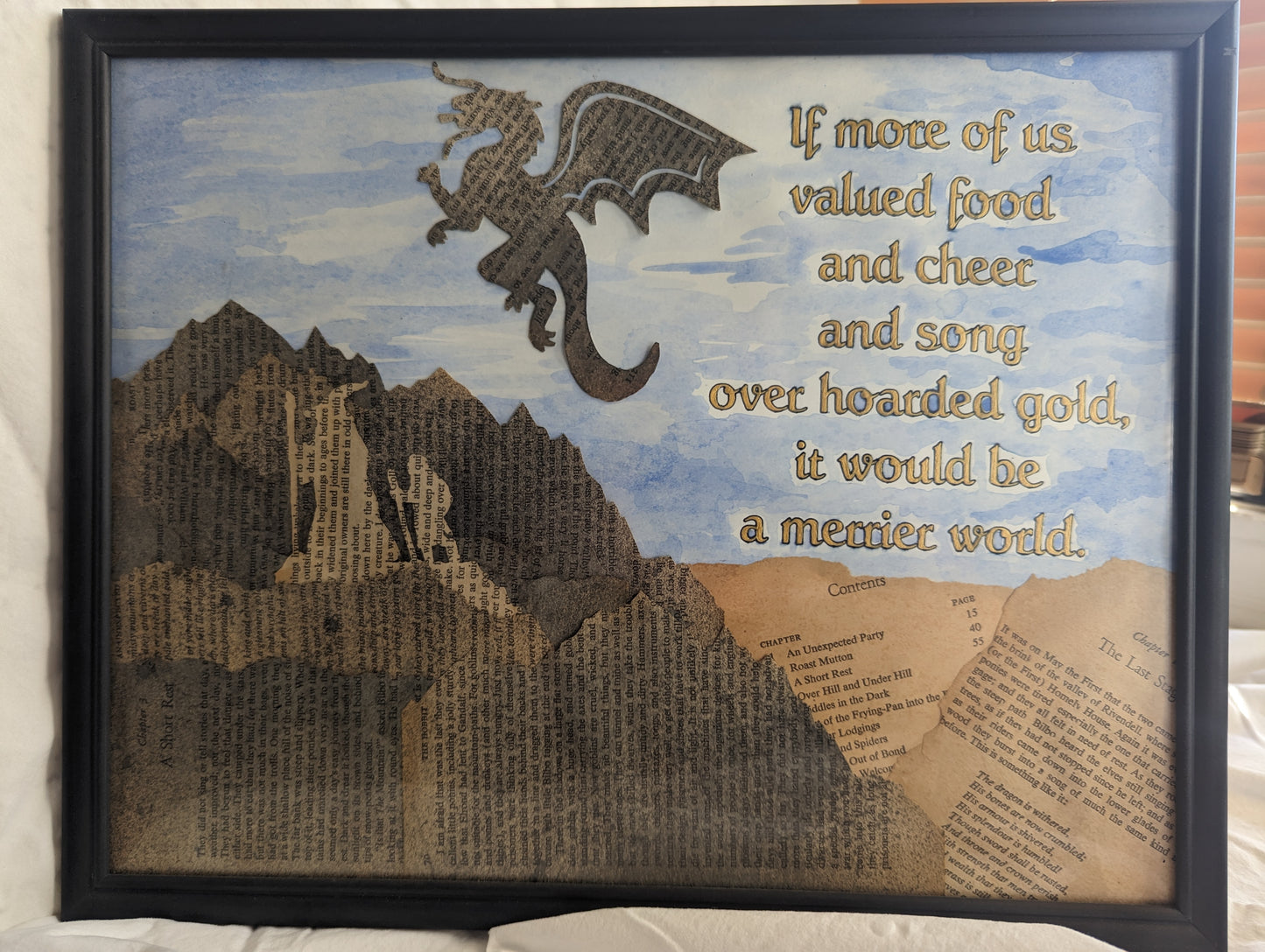 "Smaug and Hoarded Gold" is approximately 10.5x14.5 inches in a wall hanging document frame. Cut and ink stained pages from "The Hobbit" are arranged to depict Gandalf walking with Bilbo behind him through the mountains with Smaug the dragon hovering overhead. The quote, "If more of us valued food and cheer and song over hoarded gold, the world would be a merrier place." is hand painted in gold acrylic on a blue watercolor painted background.